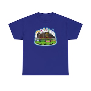 Puede incluir: Una camiseta azul real con un gráfico de las Tortugas Ninja. El diseño incluye las caras de las cuatro tortugas, un edificio y el texto "Turtles in the Clouds". El diseño es de estilo caricatura.