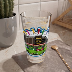 Puede incluir: Un vaso transparente lleno de hielo y líquido, con un gráfico colorido de las Tortugas Ninja. El diseño incluye el texto "Turtles in the Clouds" y "Seven Radical Evenings".
