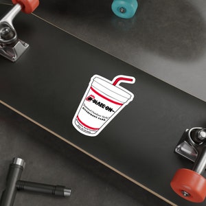 Puede incluir: Una pegatina blanca con forma de vaso con una pajita roja. La taza tiene un diseño a rayas rojas y blancas y las palabras "BLAZE-ON" y "Flammed Outdoor Style" están impresas en la taza. La pegatina también incluye el texto "Riverfront Park" y "Wilmington, North Carolina July 16-19-2015".