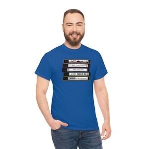 Puede incluir: Camiseta azul real con una pila de cintas VHS retro de varios colores y títulos. La camiseta está hecha de un material suave.