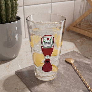 Puede incluir: Un vaso transparente lleno de hielo y rodajas de limón, con un gráfico de una botella de ketchup con el texto "Organic Fish Sauce" y "Henriettaz Est. 1983". Una cuchara de bar dorada descansa sobre la encimera.