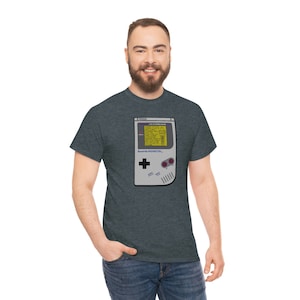 Puede incluir: Camiseta gris oscuro con un gráfico de consola de juegos portátil retro. El diseño incluye una pantalla con texto, un panel direccional y botones. El texto "Seattle Washington" está impreso debajo.