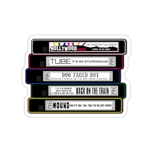 Puede incluir: Una pegatina con un diseño de pila de cintas VHS. Cada cinta tiene un color diferente y texto, incluyendo "Hollywood Bowl", "Tube", "Dog Faced Boy", "Back on the Train" y "Mound". La pegatina tiene un borde blanco.