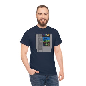 Puede incluir: Camiseta azul marino con un diseño gráfico colorido que se asemeja a un cartucho de videojuego retro. El diseño incluye el texto "PHISH" y otros gráficos. La camiseta está hecha de un material suave y tiene un cuello redondo clásico.