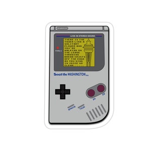 Puede incluir: Una pegatina gris con forma de consola de juegos portátil retro. La pantalla muestra una imagen amarilla del Space Needle de Seattle y texto. El texto incluye "Seattle WASHINGTON PH", "LIVE IN STEREO SOUND" y "TYPE 2".