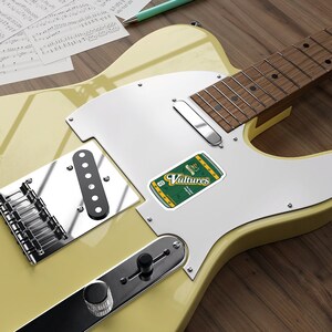 May include: A yellow electric guitar with a silver pickguard and a sticker of a green can with the text "Vultures" on it.