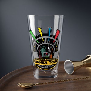 Puede incluir: Un vaso de pinta transparente con un gráfico colorido. El gráfico presenta un diseño de cabina estilizado con guitarras y el texto "SUMMER TOUR A WAVE OF HOPE EPISODE XL". El vaso está sobre una bandeja de madera con una cuchara y un jigger dorados.