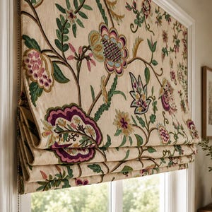 Puede incluir: Un estor romano beige con un intrincado patrón de bordado floral multicolor. El diseño incluye grandes flores estilizadas y enredaderas en tonos morado intenso, magenta, verde azulado, verde y dorado que cubren la superficie de la tela.