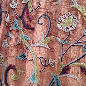 May include: Close-up of a peach fabric with detailed floral embroidery. The embroidery uses light blue, green, yellow, and burgundy threads, creating a textured design. The fabric has a soft, velvety appearance, with a rich, warm tone.