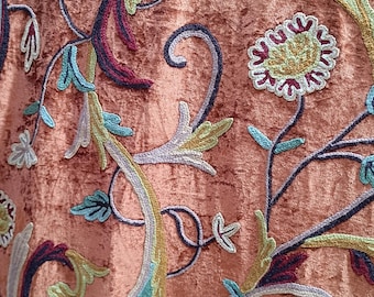 May include: Close-up of a peach-colored fabric with intricate floral embroidery. The embroidery features a variety of colors, including light blue, green, yellow, and burgundy, creating a detailed and textured design. The fabric has a soft, velvety appearance.