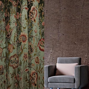 Op de afbeelding: Een groene gordijn met een bloemenpatroon in tinten rood, bruin en groen. Naast het gordijn staat een grijze fauteuil met een roze kussen. De achtergrond is een getextureerde bruine muur.
