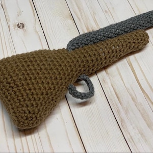 May include: A crocheted toy gun with a brown barrel and a gray handle. The toy gun is made of yarn and has a realistic design.