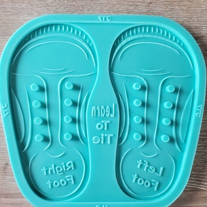 Puede incluir: Un molde de silicona turquesa para hacer dos pares de zapatos de bebé. El molde tiene dos secciones, una para el pie izquierdo y otra para el pie derecho. Cada sección tiene un diseño en relieve de un zapato de bebé con un dedo del pie, un talón y cordones. El molde también tiene las palabras "Tie the Leggin" y "Left foot" y "Right foot" inscritas en él.