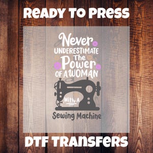 Może przedstawiać: Projekt transferu DTF z tekstem "READY TO PRESS" na górze i "DTF TRANSFERS" na dole. Projekt zawiera cytat "Never underestimate the power of a woman with a sewing machine" z grafiką maszyny do szycia.