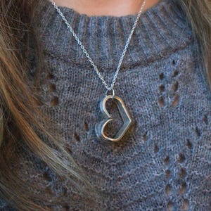 Puede incluir: Un collar con colgante de corazón plateado. El colgante tiene forma de corazón hueco, suspendido de una delicada cadena plateada. El collar se lleva sobre un jersey gris.