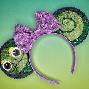 May include: Purple headband with green and gold glitter ears. One ear features a green snail with a gold spiral shell. The other ear features a green frog with large eyes.