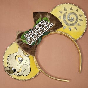 May include: Yellow and gold mouse ears headband with a Simba graphic and a sun design. The headband features a brown bow with the words "Hakuna Matata". The ears are trimmed with gold rope.