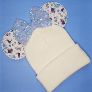 May include: White knitted beanie with a large blue sequin bow and two fuzzy ear flaps featuring a Frozen character print.