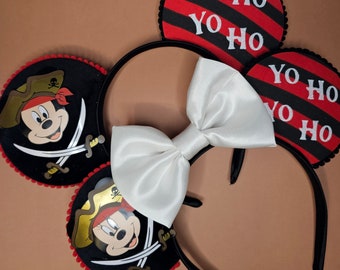 Puede incluir: Diadema negra con un lazo blanco y dos orejas de Mickey Mouse rojas y negras. Las orejas tienen un diseño de Mickey Mouse pirata con el texto "Yo Ho Yo Ho" en cada oreja.