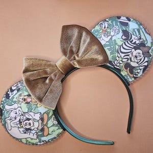 May include: A pair of brown velvet Minnie Mouse ears with a green headband. The ears are decorated with a jungle scene featuring Mickey Mouse, Donald Duck, Goofy, and other Disney characters.