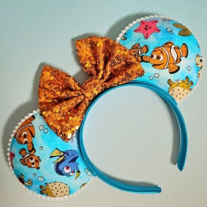 May include: A pair of blue headband mouse ears with a gold glitter bow. The ears are decorated with a Finding Nemo theme featuring characters like Dory, Marlin, and Nemo.