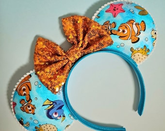 May include: A pair of blue headband mouse ears with a gold glitter bow. The ears are decorated with a Finding Nemo theme featuring characters like Dory, Marlin, and Nemo.