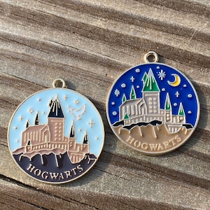 Puede incluir: Dos abalorios de metal de color dorado que representan una ilustraci&oacute;n detallada del castillo de Hogwarts. Un abalorio tiene un fondo azul con estrellas y una luna creciente. El otro abalorio tiene un fondo blanco con estrellas. Ambos abalorios tienen la palabra "HOGWARTS" escrita en la parte inferior.