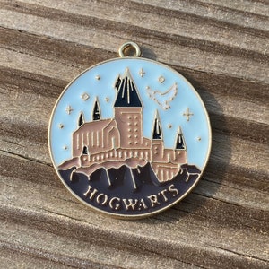 Puede incluir: Un amuleto de metal dorado con fondo de esmalte blanco que presenta una ilustraci&oacute;n detallada del castillo de Hogwarts con el texto "HOGWARTS" en letras negras.