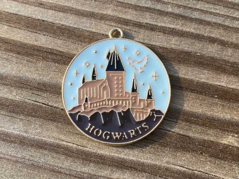 Puede incluir: Un amuleto de metal dorado con fondo de esmalte blanco que presenta una ilustraci&oacute;n detallada del castillo de Hogwarts con el texto "HOGWARTS" en letras negras.