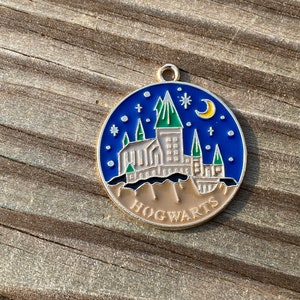 Puede incluir: Un pin de esmalte azul y dorado con una ilustraci&oacute;n detallada del castillo de Hogwarts con una luna creciente y estrellas. El pin dice "HOGWARTS" en la parte inferior.
