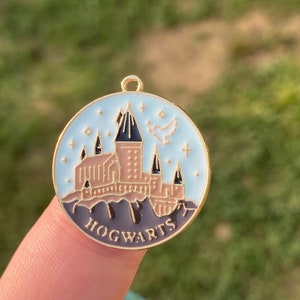 Puede incluir: Un amuleto de metal dorado con un fondo de esmalte azul que presenta una ilustraci&oacute;n detallada del castillo de Hogwarts con un p&aacute;jaro volando por encima. El amuleto tiene la palabra "HOGWARTS" escrita en letras doradas en la parte inferior.
