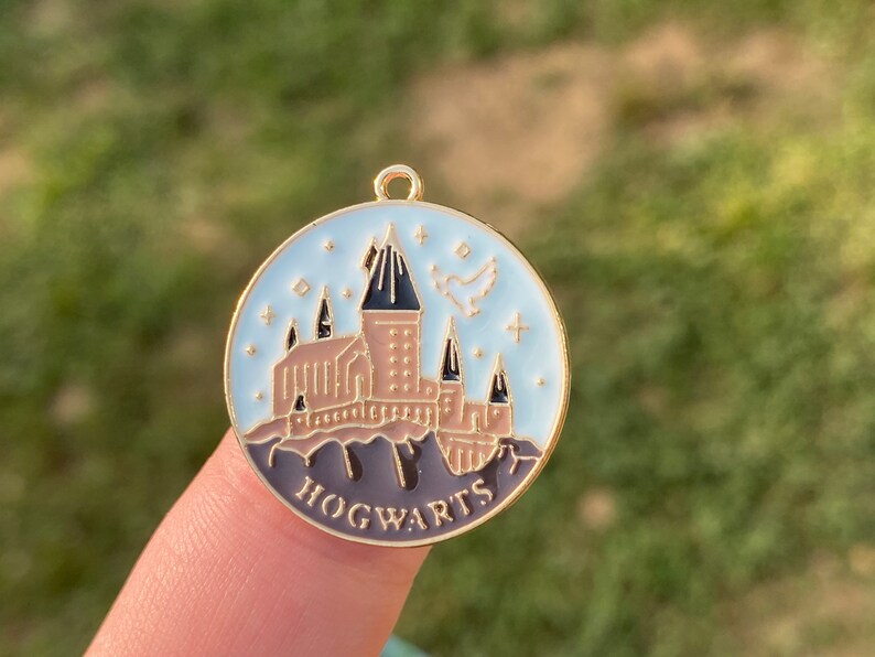 Puede incluir: Un amuleto de metal dorado con un fondo de esmalte azul que presenta una ilustraci&oacute;n detallada del castillo de Hogwarts con un p&aacute;jaro volando por encima. El amuleto tiene la palabra "HOGWARTS" escrita en letras doradas en la parte inferior.