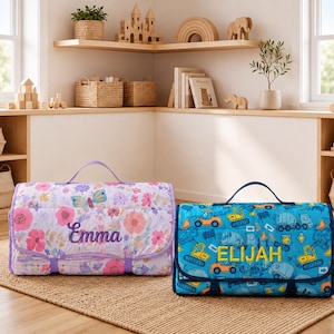 May include: Two rectangular, foldable nap mats with carrying handles. The left mat features a light purple floral pattern with a butterfly and the name Emma embroidered in purple. The right mat has a blue construction vehicle print with the name ELIJAH embroidered in yellow.