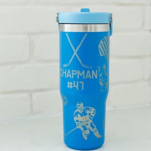 May include: A blue insulated tumbler with a light blue lid and silver accents. The tumbler features hockey-themed graphics and the name "Chapman" with the number "47". The tumbler is designed to keep beverages cold or hot.