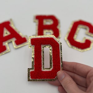 May include: Four felt letter patches in red with gold trim. The letters are A, B, C, and D. The patches are on a white background.