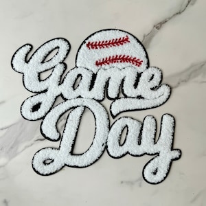 May include: A white, textured "Game Day" patch with a black border. The baseball-themed design includes a baseball with red stitching. The patch is likely for apparel or accessories, perfect for sports fans.