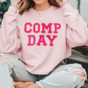 May include: A pink sweatshirt with the text "COMP DAY" in gold glitter lettering.