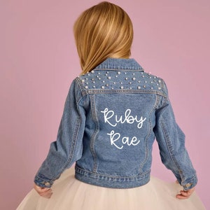 May include: A blue denim jacket with pearl embellishments on the shoulders and the name "Ruby Rae" embroidered on the back.