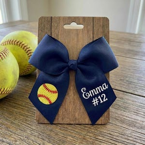May include: Navy blue softball bow with a yellow softball graphic and the name "Emma" and the number "12" embroidered in white. The bow is attached to a wooden display card. Two softballs are in the background.