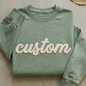 May include: A sage green sweatshirt with the word "custom" in a cream-colored, textured script across the front. The sweatshirt has a crew neck and long sleeves with ribbed cuffs. The garment is folded neatly on a neutral surface.