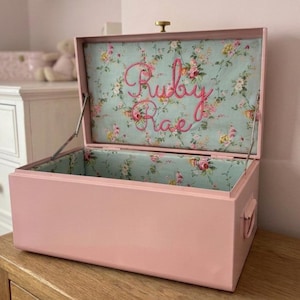 May include: A rectangular pink metal storage chest with a hinged lid. The interior is lined with light blue fabric featuring a floral pattern. The name Ruby Rae is embroidered in pink script on the inside of the lid. It has a gold knob.