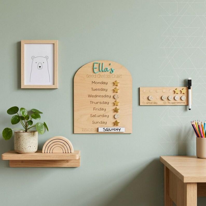 Puede incluir: Un cuadro de buenas elecciones de madera con el nombre "Ella's" en escritura turquesa. El cuadro incluye los d&iacute;as de la semana, iconos de estrellas y emoticonos, y la palabra "SQUISHY" en una etiqueta blanca. Tambi&eacute;n se ve una impresi&oacute;n de oso enmarcada, un arco&iacute;ris de madera y una planta peque&ntilde;a.