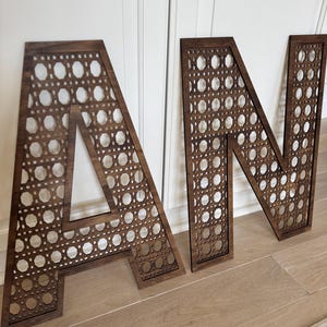 K&ouml;nnte beinhalten: Holzbuchstaben 'A' und 'N' mit dunkelbraunem Finish. Die Buchstaben haben ein gewebtes Rohrdesign innerhalb der Buchstabenformen. Die Buchstaben werden auf einem hellen Holzboden mit wei&szlig;em Hintergrund angezeigt.