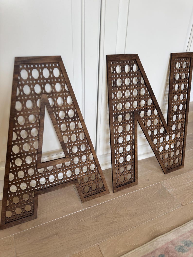 K&ouml;nnte beinhalten: Holzbuchstaben 'A' und 'N' mit dunkelbraunem Finish. Die Buchstaben haben ein gewebtes Rohrdesign innerhalb der Buchstabenformen. Die Buchstaben werden auf einem hellen Holzboden mit wei&szlig;em Hintergrund angezeigt.