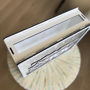 Peut inclure: Un cadre en bois blanc et noir avec le mot "MAKE" visible. Le cadre est ouvert, r&eacute;v&eacute;lant une surface r&eacute;fl&eacute;chissante. Il repose sur un tabouret rond de couleur claire avec un motif ray&eacute;.