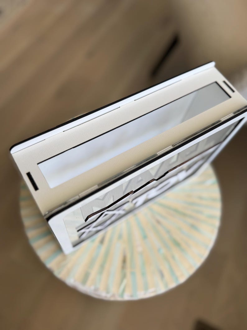Peut inclure: Un cadre en bois blanc et noir avec le mot "MAKE" visible. Le cadre est ouvert, r&eacute;v&eacute;lant une surface r&eacute;fl&eacute;chissante. Il repose sur un tabouret rond de couleur claire avec un motif ray&eacute;.