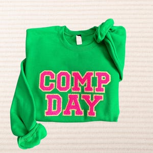 May include: Green sweatshirt with the words "COMP DAY" in pink glitter letters.