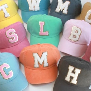 May include: A collection of ten baseball caps in various colors, each featuring a large, white felt letter on the front. The letters are A, B, C, H, J, L, M, S, T, and W.