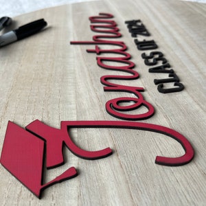 May include: A wooden sign with the words "Congratulations Class of 2024" in black lettering. A red graduation cap is in the lower left corner of the sign.