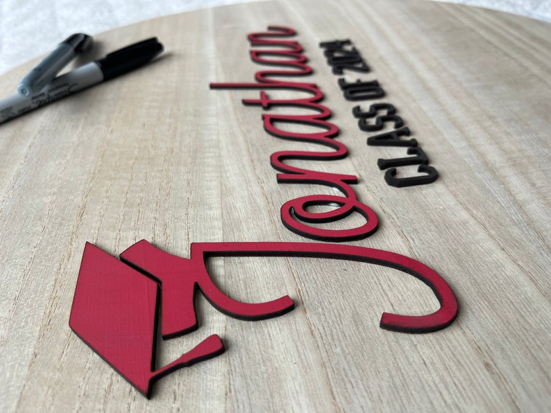 May include: A wooden sign with the words "Congratulations Class of 2024" in black lettering. A red graduation cap is in the lower left corner of the sign.